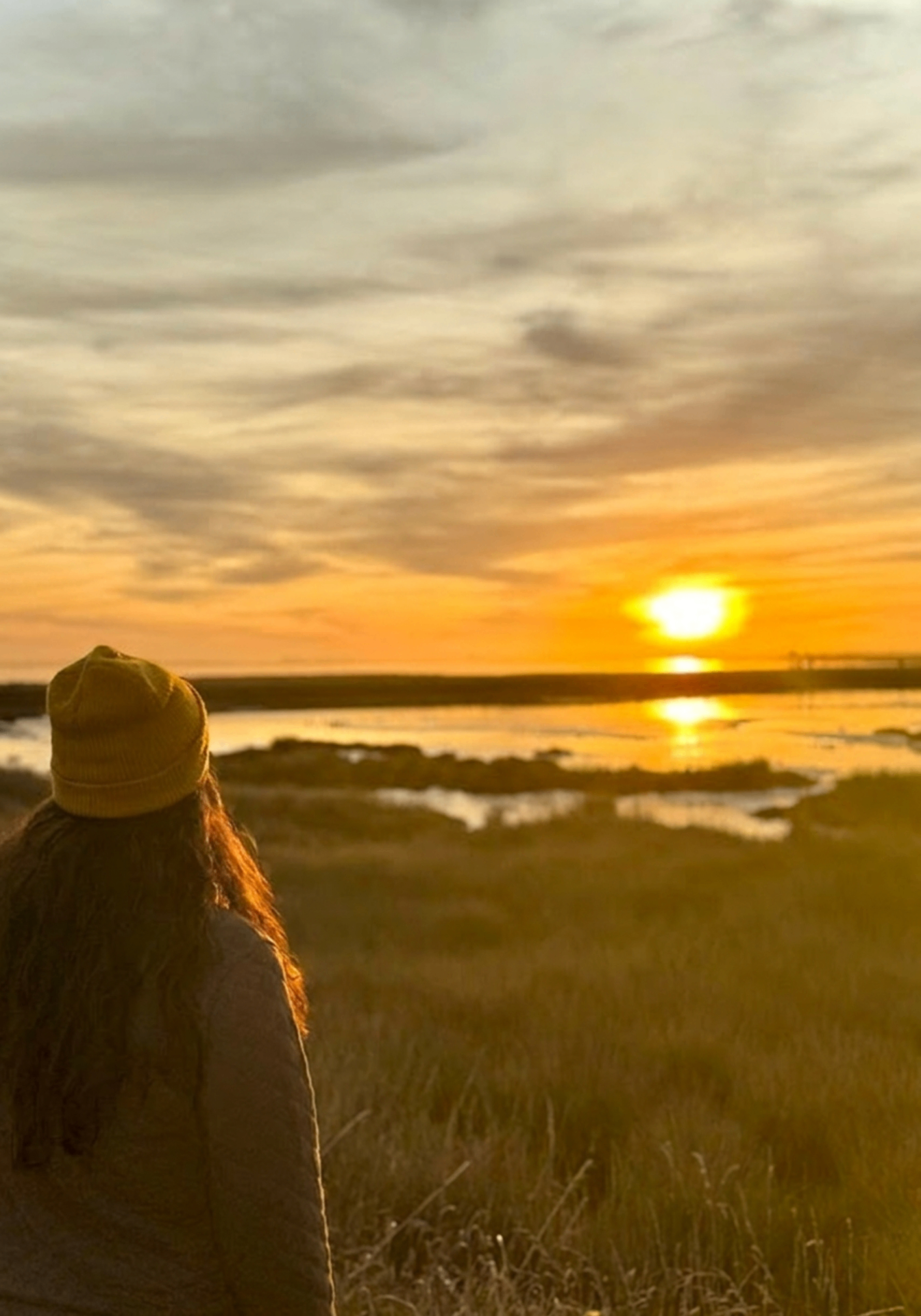 Woman in a yellow beanie facing a golden sunset over a coastal marsh.